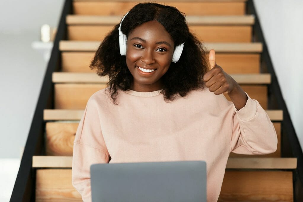 Black Female Using Laptop Gesturing Thumbs-Up Sitting On Steps Indoor