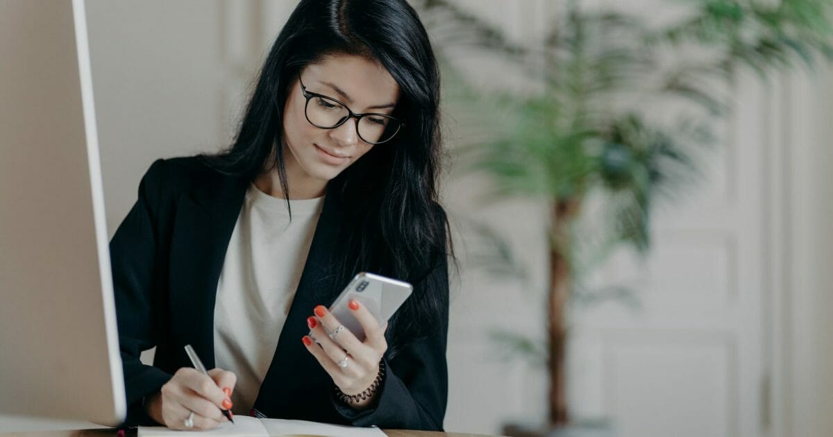 Woman with dark hair, concentrated into mobile phone, searches necessary information in internet